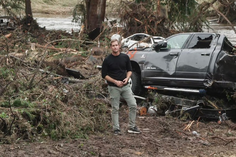 Morgan Chesky stands in the forest among flood debris and a damaged vehicle