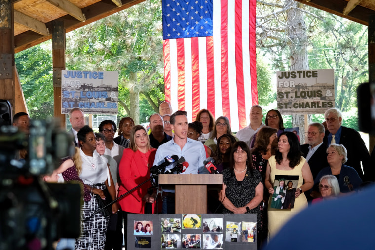 Josh Hawley speaks at a podium outside, people surround him, and American flag hangs behind him
