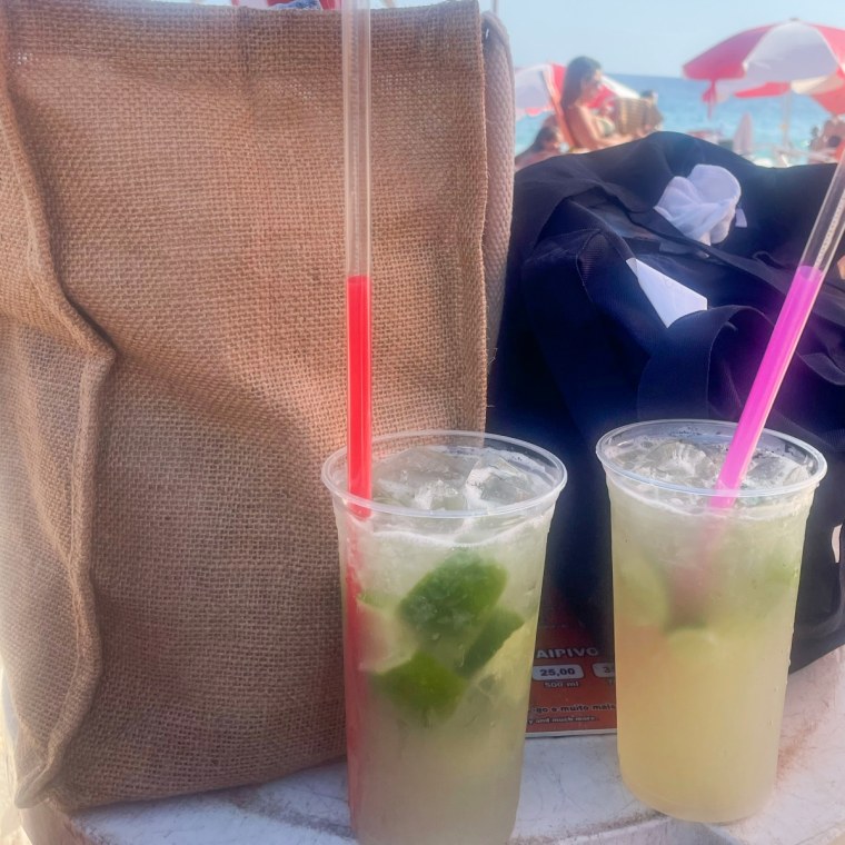 The Muji Jute bag on a white table beside a black tote bag, with two lime drinks in front and a beach in the background.