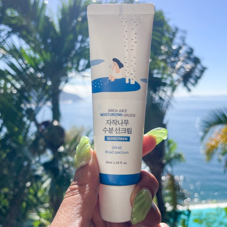 A left hand with green nails holding the Round Lab sunscreen in front of an ocean background.