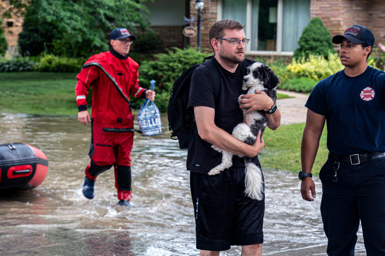 A resident carries a dog in floodwaters as first responders help.