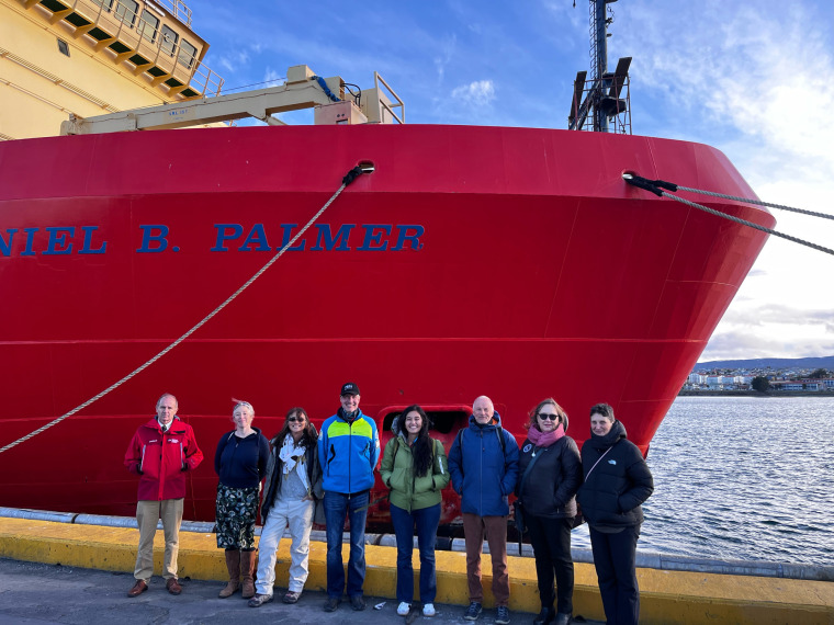 A row of eight people stand in front of a large red ship