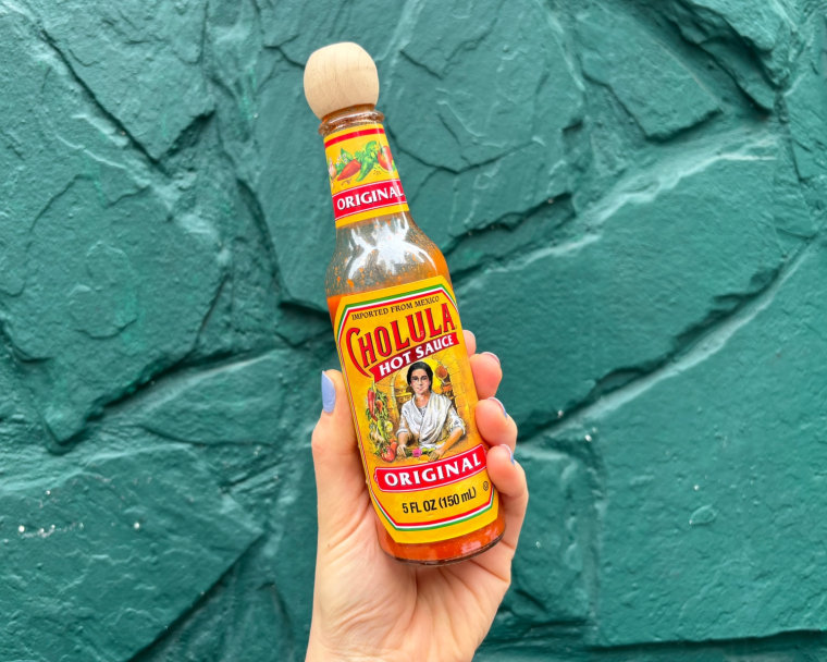 A person’s hand holding a bottle of Cholula in front of a teal wall.