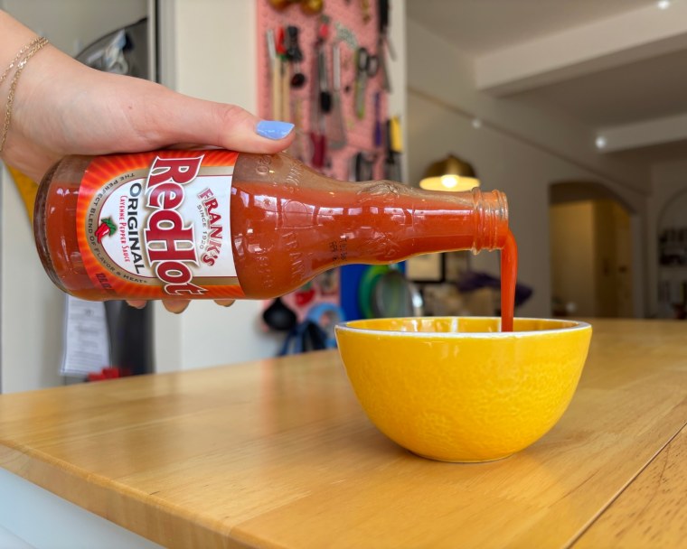 A hand pouring Frank’s Red Hot Original Hot Sauce into a small yellow bowl.