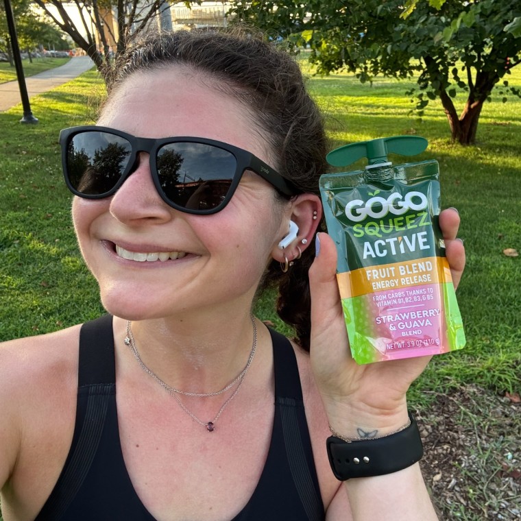 A woman holding a GoGo Squeez Active Fruit Blend Energy Release pouch outside next to her face.