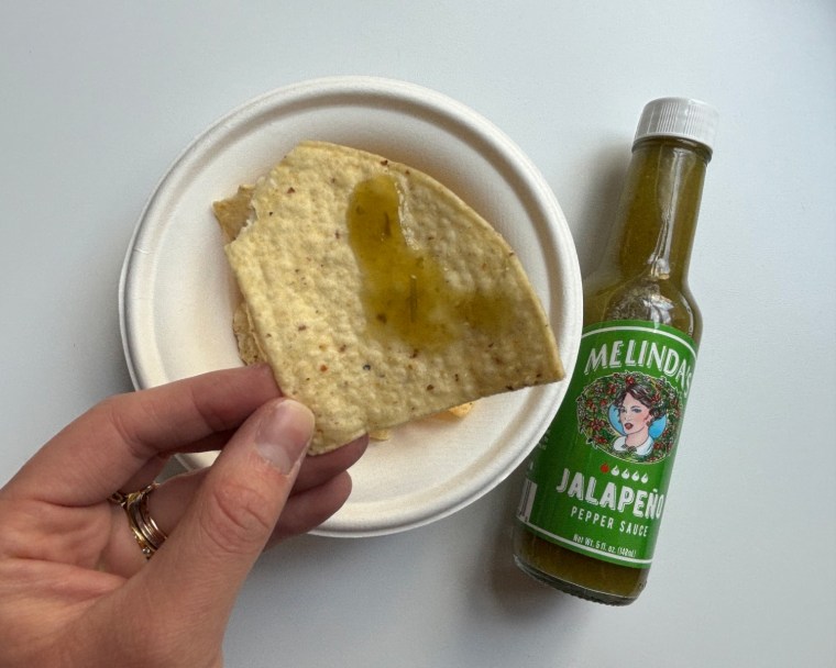 A hand holding a tortilla chip with Melinda’s Jalapeño Pepper Hot Sauce on it and a bottle of the sauce in the background.