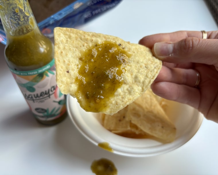 A hand holding a tortilla chip with Pisqueya’s Medium Heat Hot Sauce on it and a bottle of the sauce in the background.