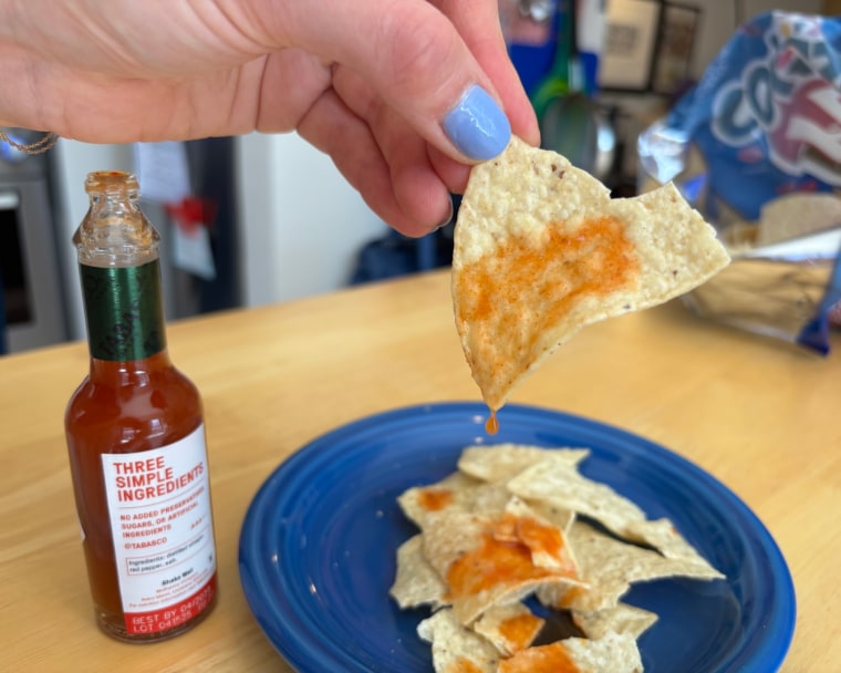 A hand shaking a few drops of Tabasco Original Red Pepper Hot Sauce onto a tortilla chip.