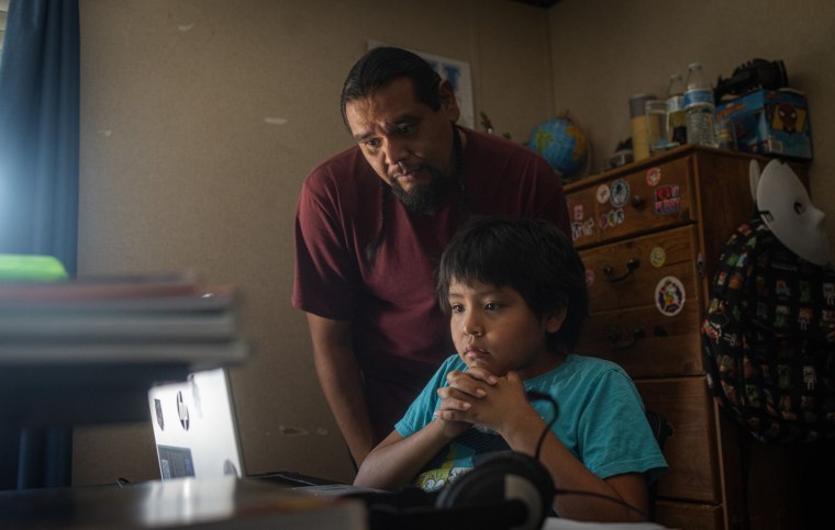 Evan Torres looks over Isaiah's shoulder while they look at a laptop inside of a bedroom