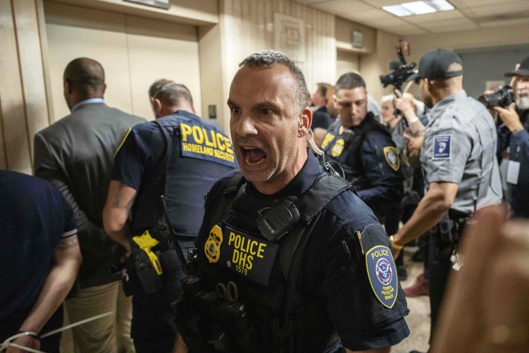 Federal agent in the centre of the frame, with elected officials being detained in the background