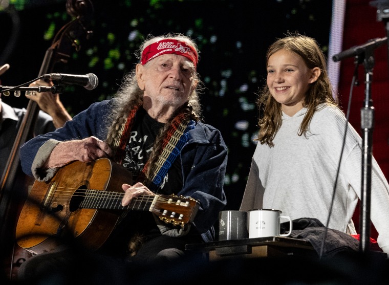 Willie Nelson, left, sits on stage holding a guitar next to Charlie Marie Selman