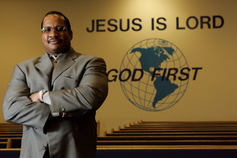 Patrick Wooden stands in front of pews in a church, text on the wall behind him reads "Jesus is Lord, God First"