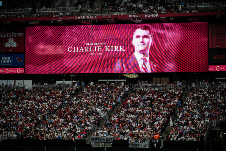 People sit in bleachers at the Charlie Kirk memorial as a billboard showing Charlie Kirk on the screen behind them