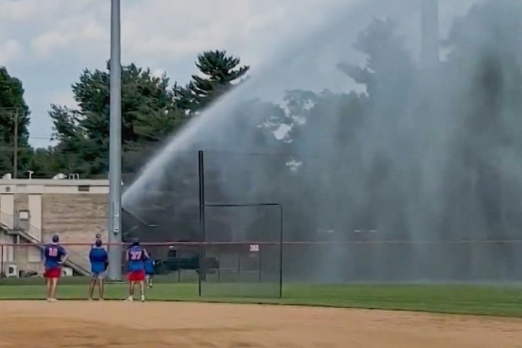 Three baseball players watch as water is sprayed onto the baseball field