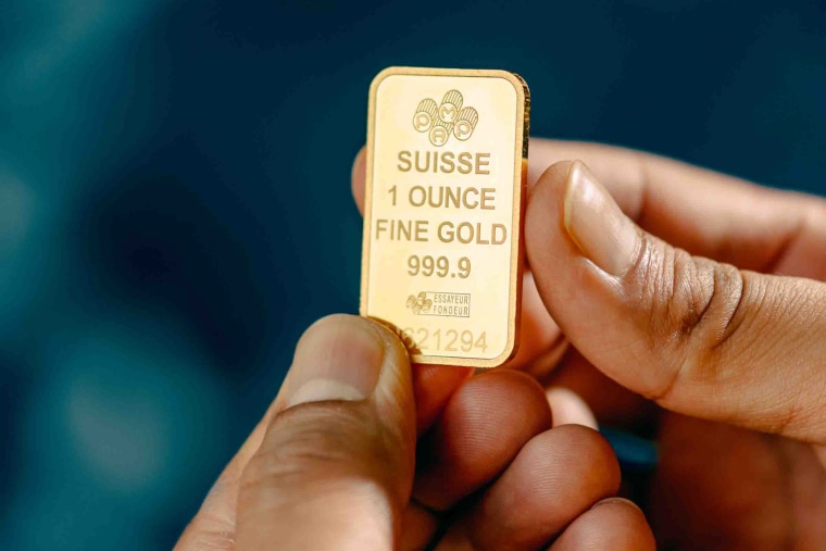 Two hands hold a small gold bar with imprinted words that read "Suisse 1 Ounce Fine Gold 999.9"