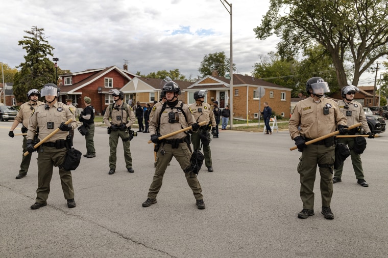 Illinois State police officers armed with wooden batons stand guard as protesters gather outside the Broadview ICE processing facility