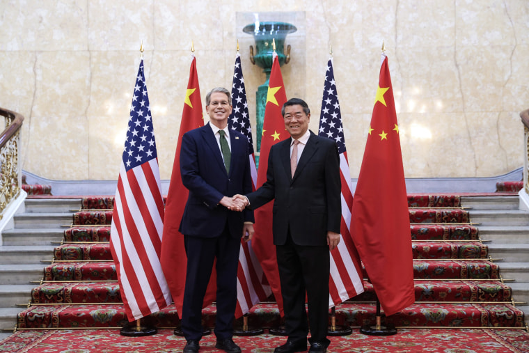 Scott Bessent shakes hands with He Lifeng while posing at the bottom of a stairwell and smiling, American and Chinese flags stationed behind them