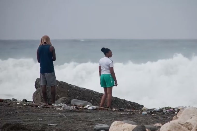 Two people watch waves crash by the rocks outside