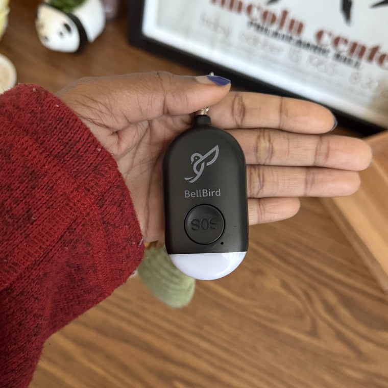 A left hand holding a black and white Bellbird Safety Alarm in front of a brown, wooden desk.