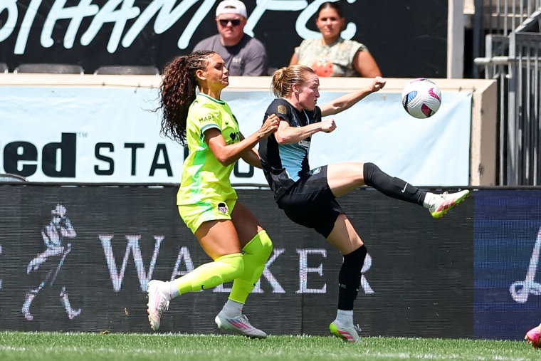 Trinity Rodman, à esquerda, corre atrás de Emily Sonnett enquanto Sonnett acerta uma bola de futebol no ar no campo.