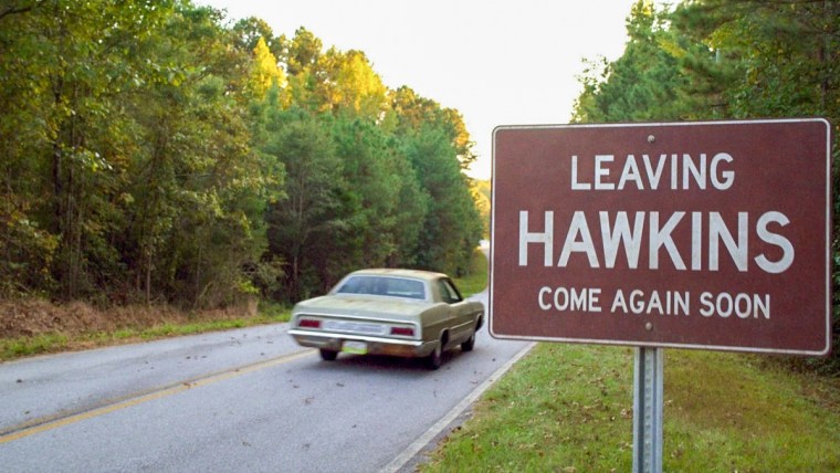 A car on a road passes by a red sign with white text that reads "Leaving Hawkins Come Again Soon"
