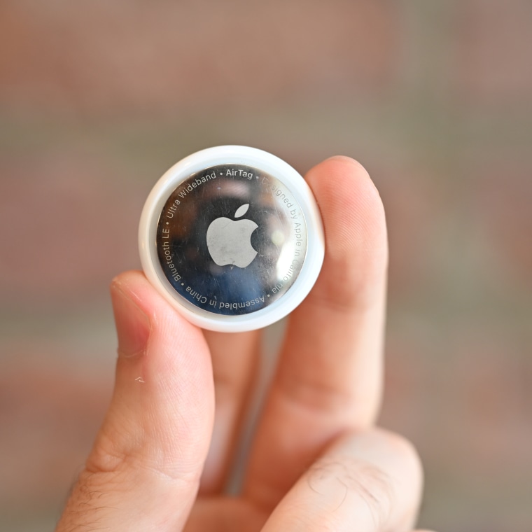 The writer holding an Apple AirTag in between his fingers against a brick background.