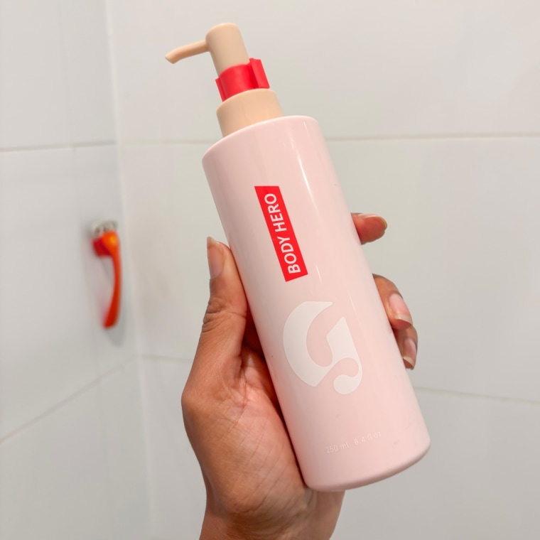 A hand holding a pink and red body wash bottle with a bathtub background.