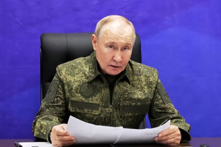Russian President Vladimir Putin sat holding a papers against a purple background