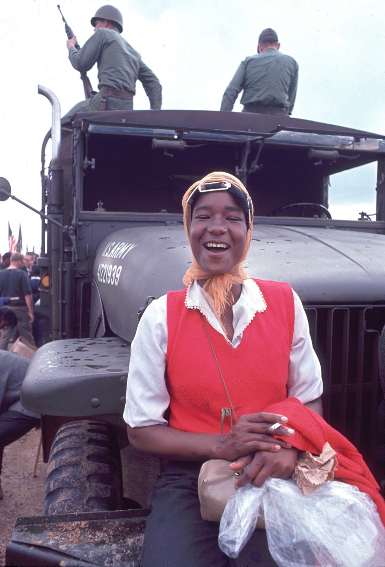 Doris Wilson smokes a cigarette outside up against an army vehicle with two soliders on top outside