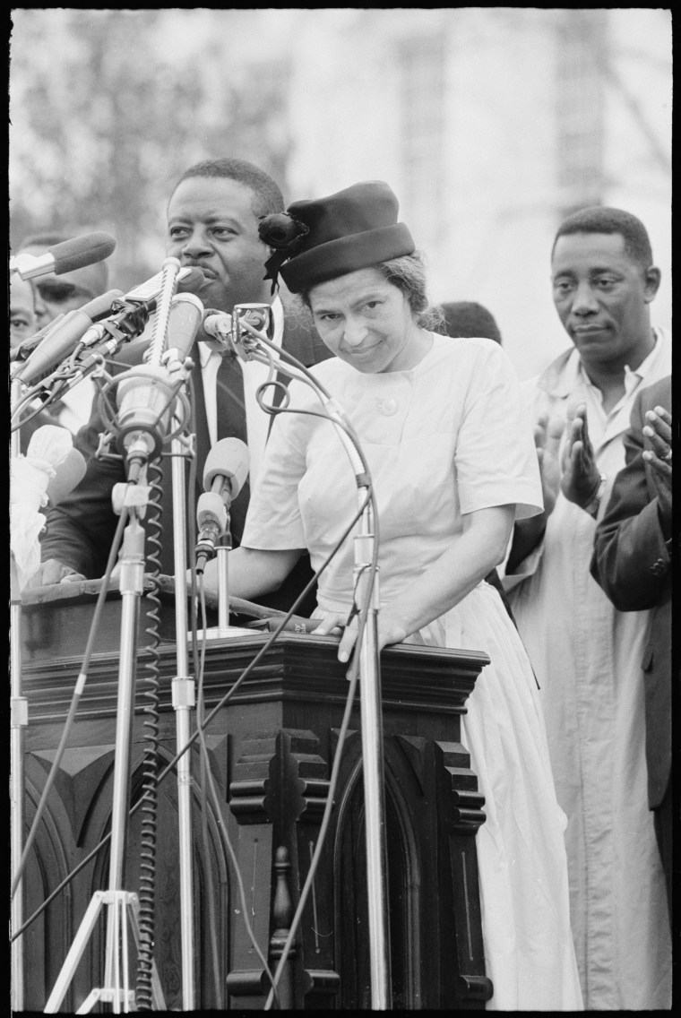 Rosa Parks speaks at a podium outside as people stand behind her