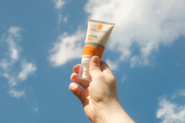 A hand holds tone bottle of sunscreen against a blue sky with clouds