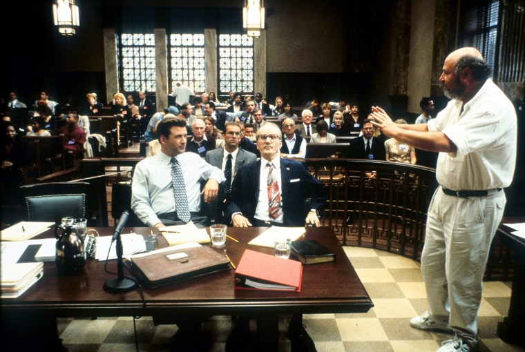 Alec Baldwin, left, and James Woods sit at a table in a courtroom, Rob Reiner directs them on the right, people sit behind them in the pews