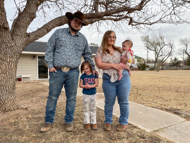 A father, mother, and their two small daughters stand outside of a house and pose for a family photo