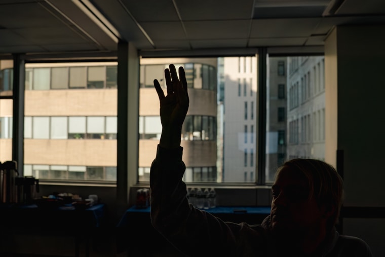 The silhouette of a raised hand can be seen in a classroom, in the back of the room is a large set of windows facing a building during the daytime