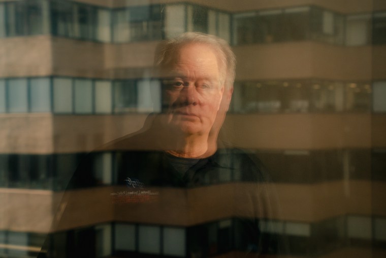 Rob Weil stands for a portrait, the image is of his reflection in a window facing a building