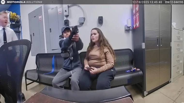 A man holds out a gun, attempting to shoot it, while seated on a couch next to a woman inside of a room. On the left, a guard yells and runs towards him