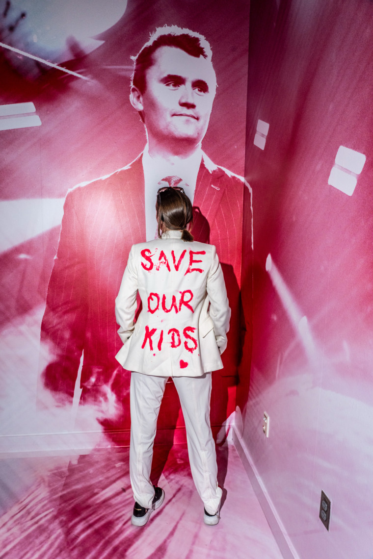 A person is turned towards a wall, showing the back of their white suit jacket that reads "Save Our Kids" in red paint, the wall has a picture of Charlie Kirk on it