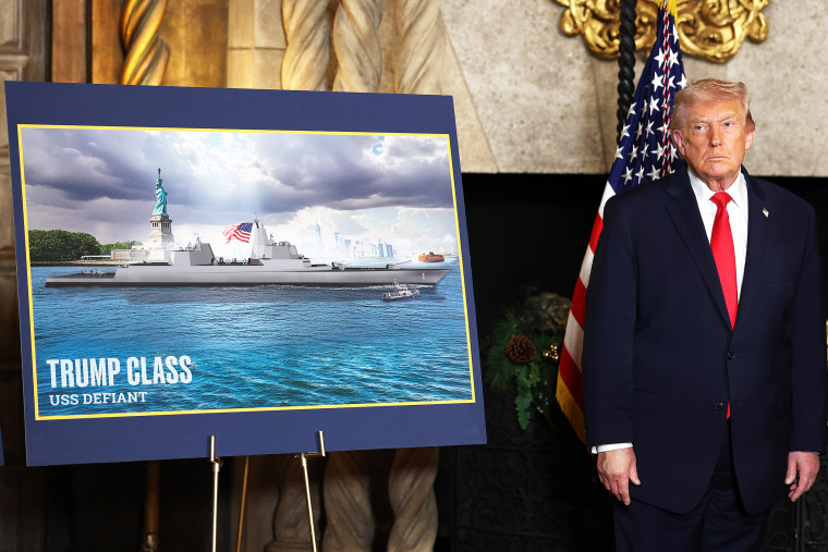 Donald Trump stands next to a signboard with an image of a battleship inside a room at Mar-a-Lago.