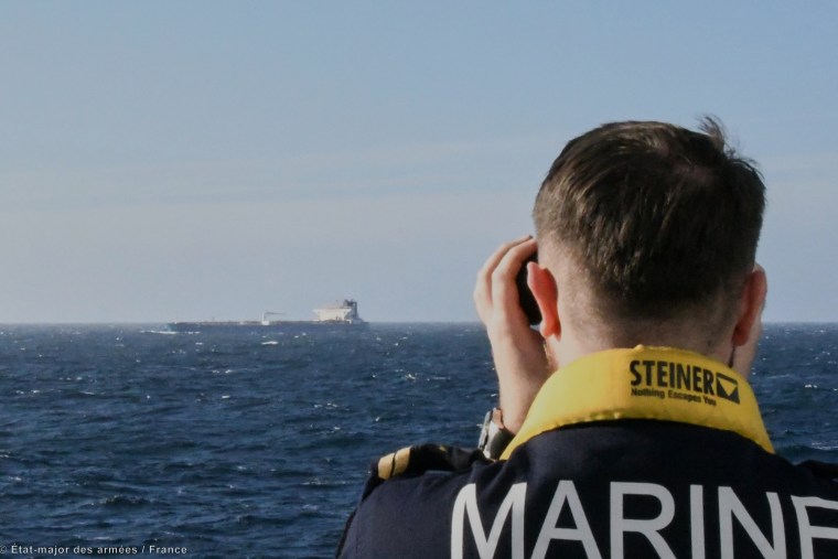 The back of a person's head can be seen, they are looking into binoculars over the ocean at an oil tanker