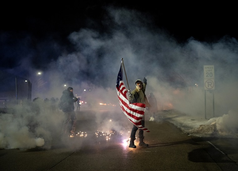 A protester holds an American flag while surrounded by smoke and flares.