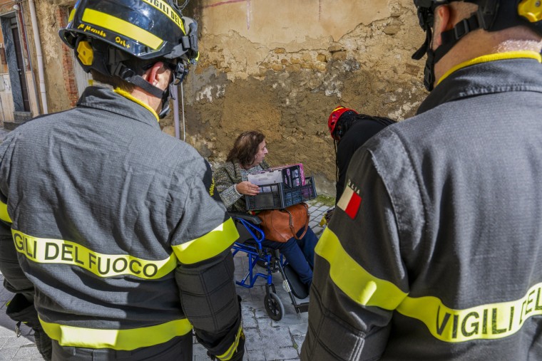 A disabled woman in a wheelchair is assisted by firefighters while carrying personal belongings on her lap.