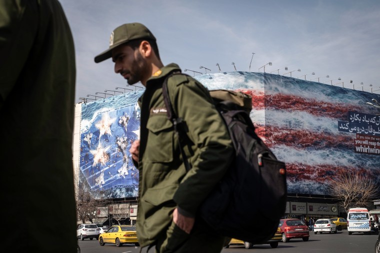 A soldier wearing green fatigues walks in front of a large building wrapped in a graphic bearing the stars and stripes of the American flag.
