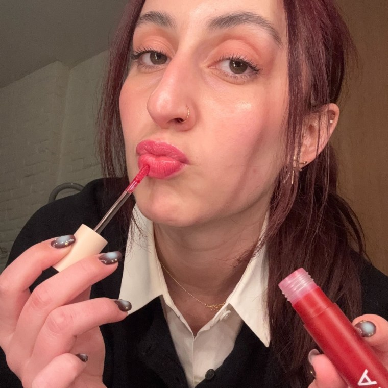 Woman applying red lip gloss to puckered lips while holding a gloss tube.