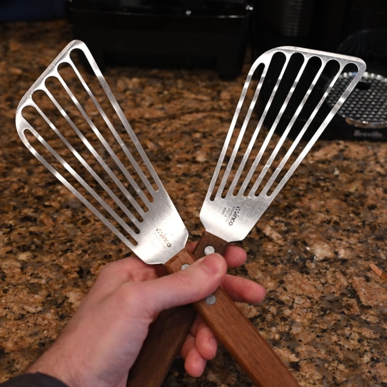 A close-up of someone holding two stainless steel spatulas in one hand over a kitchen counter top.