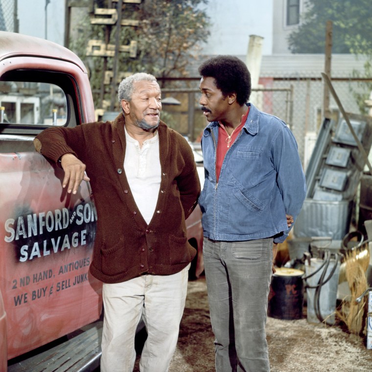 Red Foxx, left, and Demond Wilson stand next to a red truck that reads "Sanford and Son Salvage" on the driver's side door