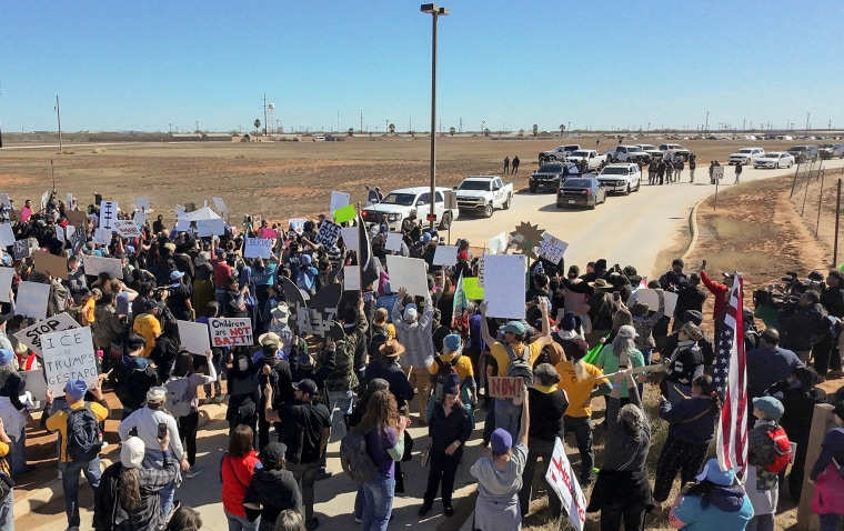 Hundreds of protesters holding signs stand in the middle of a road in a dusty landscape. Law enforcement vehicles and officers on foot stand opposite them in the distance.