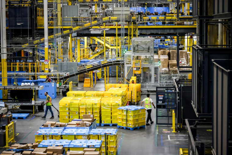A wide view of the distribution floor of an Amazon warehouse, workers move stack of packages near sorting machines