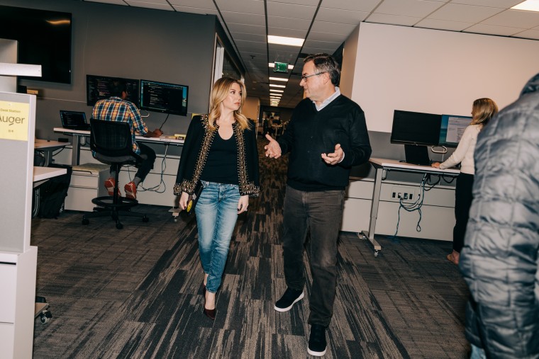 Leigh Anne Clark and Dave Clark walk in the middle of an office while talking to each other, workers are at computers around them