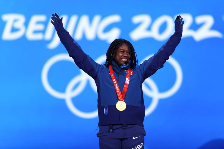 Erin Jackson mengangkat tangannya untuk merayakan, mengenakan kalung medali emas di depan latar belakang biru dengan teks putih bertuliskan: Beijing 2022"