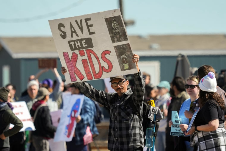 A man holding a sign reading SAVE THE KIDS stands among a crowd of fellow protesters.